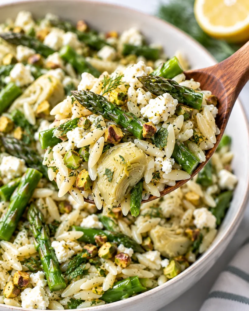 Close-up of orzo salad with asparagus, artichoke hearts, feta, pistachios, and herbs being lifted from a modern ceramic bowl on a bright white marble surface, showing glossy pasta and crisp green asparagus in sharp focus.