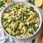 Orzo salad with asparagus, artichoke hearts, feta, pistachios, and herbs served in a modern ceramic bowl on a bright white marble surface, with glossy pasta, crisp green asparagus, creamy feta, and fresh lemony texture.