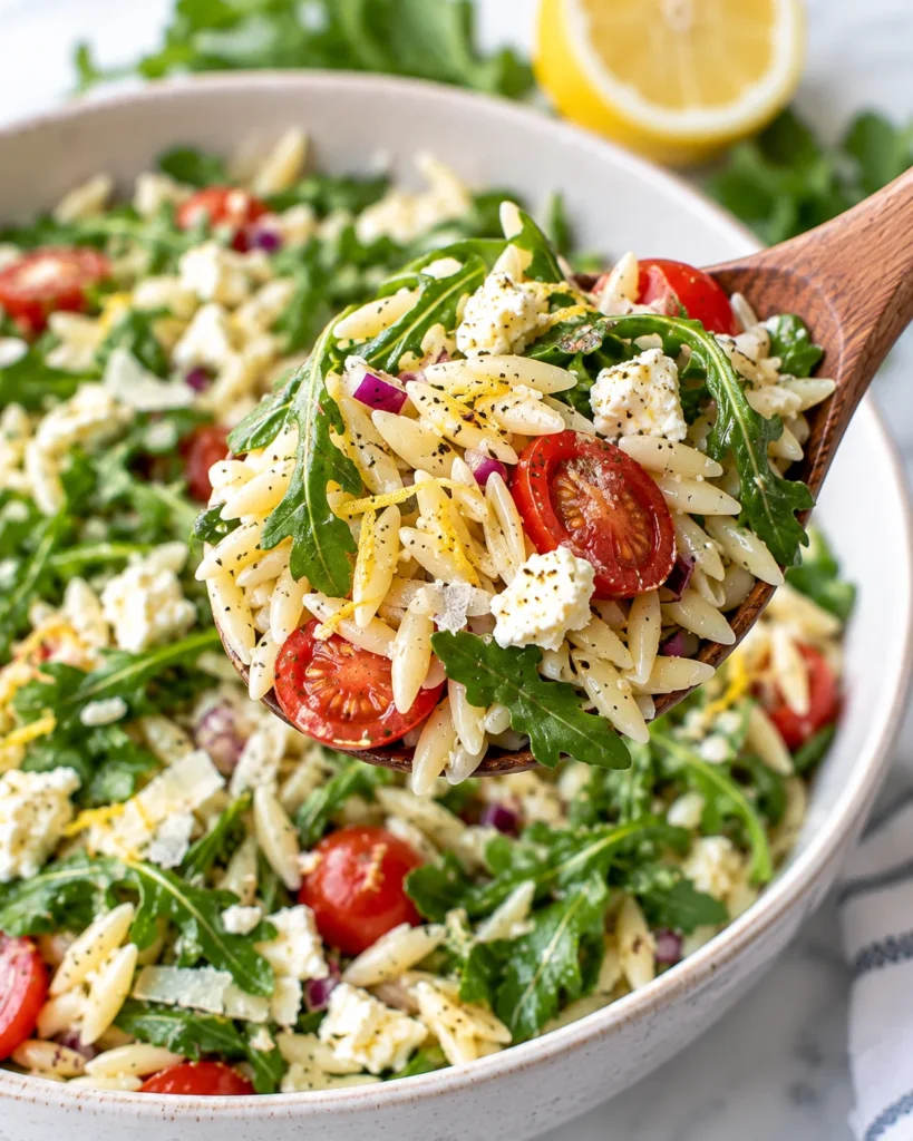 Close-up of lemon arugula orzo salad lifted on a spoon from a modern ceramic bowl on a bright white marble surface, showing glossy orzo, peppery greens, feta, chickpeas, toasted pine nuts, and lemony dressing.