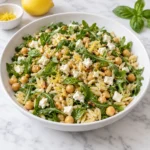 Lemon arugula orzo salad with chickpeas, crumbled feta, toasted pine nuts, basil, and lemon zest served in a modern white ceramic bowl on a bright white marble surface, showing glossy orzo and fresh peppery greens.