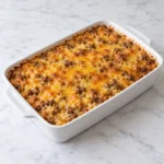 Close-up of cheesy ground beef and rice casserole being lifted from a white ceramic baking dish, revealing tender rice, browned beef, melted golden cheese, and a freshly scooped-out serving area on a bright white marble surface.
