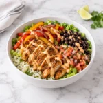 Chicken fajita bowl in a modern white ceramic bowl on bright white marble, with cilantro lime rice, seasoned sliced chicken, sautéed bell peppers and red onion, black beans, lettuce, pico de gallo, and creamy chipotle aioli.