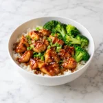 Teriyaki chicken rice bowl in a modern white ceramic bowl on bright white marble, with glossy glazed chicken, fluffy rice, broccoli, sesame seeds, and green onions.