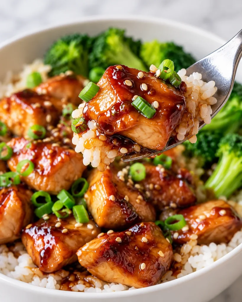 Close-up of glossy teriyaki chicken and rice lifted on a fork above the same white ceramic bowl, showing the sticky sauce and tender chicken texture against the bright white marble setting.