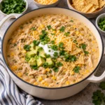 Creamy white chicken chili with shredded chicken, white beans, corn, green chiles, cheese, sour cream, avocado, cilantro, scallions, and tortilla chips served in a cream-colored Dutch oven on a bright white marble-style surface, showing a thick homemade texture.
