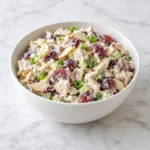 Creamy chicken salad with grapes, celery, sliced almonds, green onions, and cranberries in a white ceramic bowl on white marble.