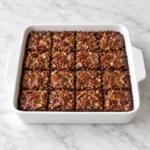 Gooey pecan pie brownies with glossy pecan topping, cut into squares in a white ceramic baking dish on white marble.