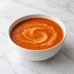 Creamy roasted tomato soup with a swirl of cream in a white ceramic bowl on white marble.