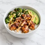 Sticky honey soy chicken, rice, broccoli, avocado, and spicy mayo in a white ceramic bowl on white marble.